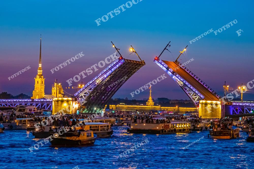 На снимке запечатлен момент разведения Дворцового моста в Санкт-Петербурге. На фоне видна Петропавловская крепость, окруженная огнями белой ночи. Нева полна катеров и лодок, из которых жители и гости города наблюдают это зрелище. Летний вечер с ярким небом и архитектурной подсветкой создает атмосферу праздника.